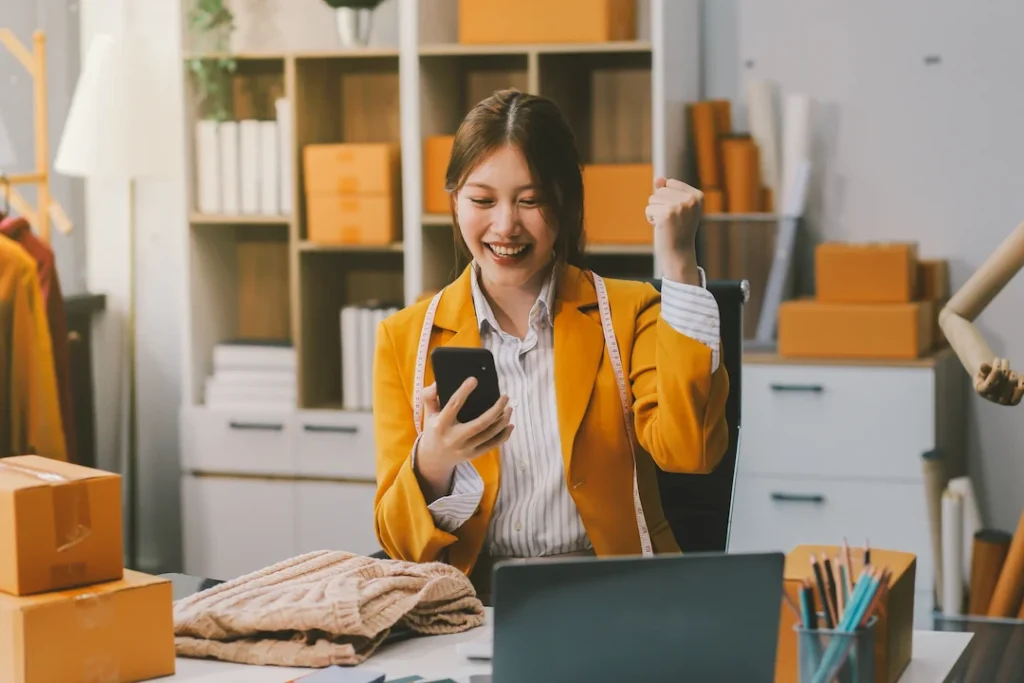 Retrato de creadora de contenido joven y carismática celebrando con su smartphone entre cajas de productos, representando talento para marcas de e-commerce en Matchis.